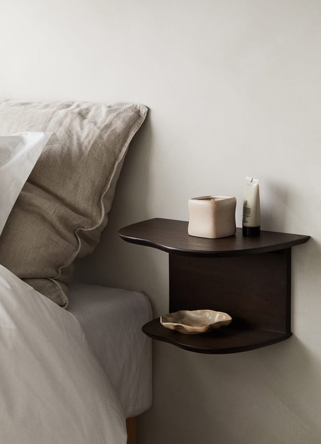 A minimal bedside setup features a dark wooden floating shelf holding high street homeware—a small beige vase, a tube, and a wavy dish. Next to the shelf is a bed with neutral bedlinen and a light cushion against a pale wall.