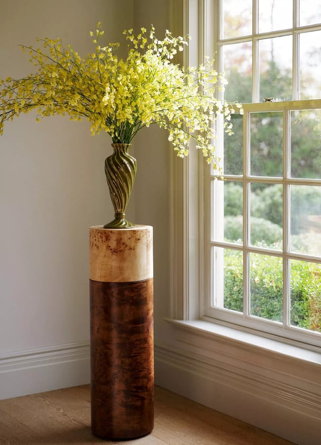 A tall brown and beige burr wood pedestal stands by a window, topped with a decorative vase of yellow flowers, with sunlight streaming in and greenery visible outside.