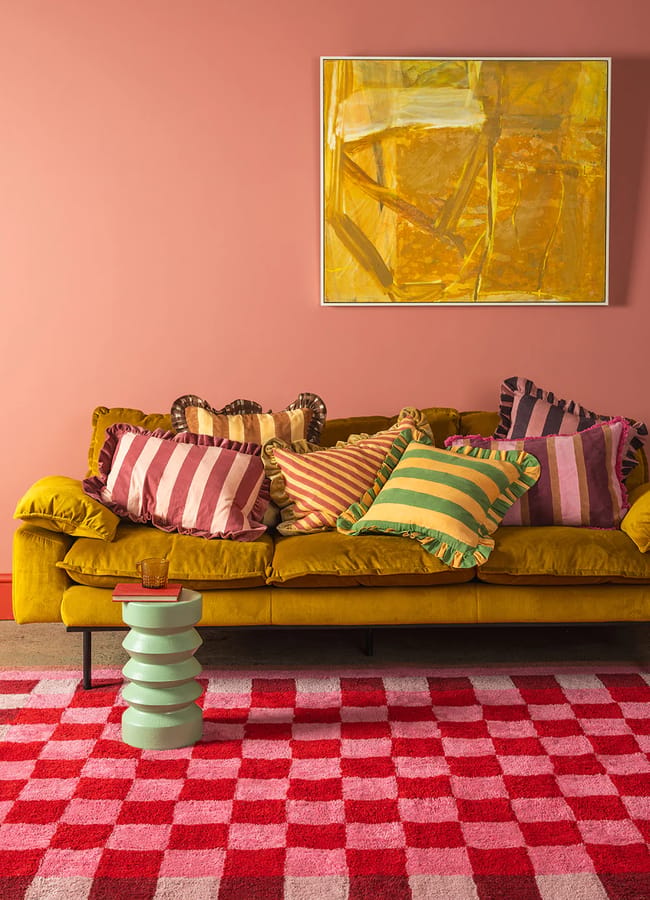 A mustard-yellow sofa with colourful striped pillows sits in front of a pink wall with a yellow abstract painting, highlighting the lively charm of red and pink interiors. A chequered rug and a light green, wavy side table complete the scene.