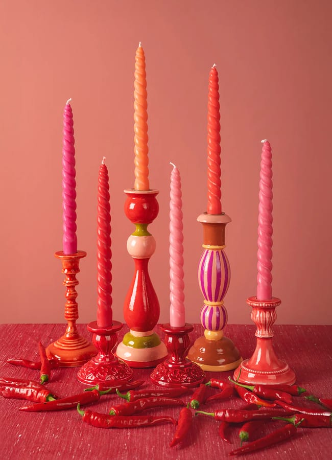 A group of pink and red twisted candles in decorative holders sit on a red surface with scattered red chilli peppers, creating a striking accent for red and pink interiors against a matching pink-red background.