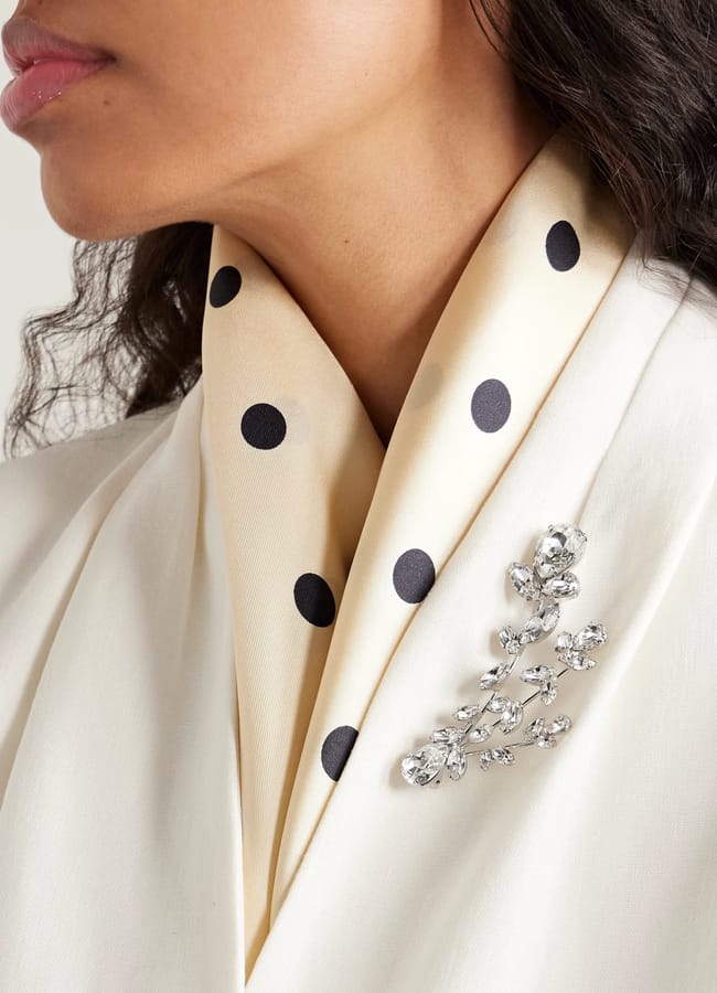 A woman wearing a cream-coloured polka dot scarf and white blazer, adorned with a sparkling silver and crystal branch-shaped brooch—a standout fashion accessory that elevates her style.