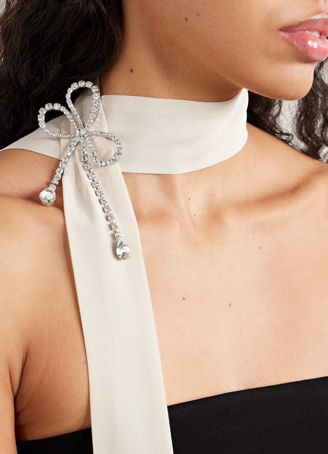 A woman wears a light cream scarf around her neck, fastened with a sparkling rhinestone bow brooch—a stylish fashion accessory that transforms her look. She has wavy dark hair and is dressed in a black, strapless top.
