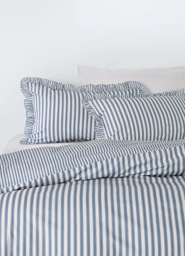 A neatly made bed features blue and white vertical striped bedding with matching pillowcases, capturing a high street homewares look. The ruffled pillowcases, plain white pillows, and white wall create an effortlessly stylish bedroom setting.