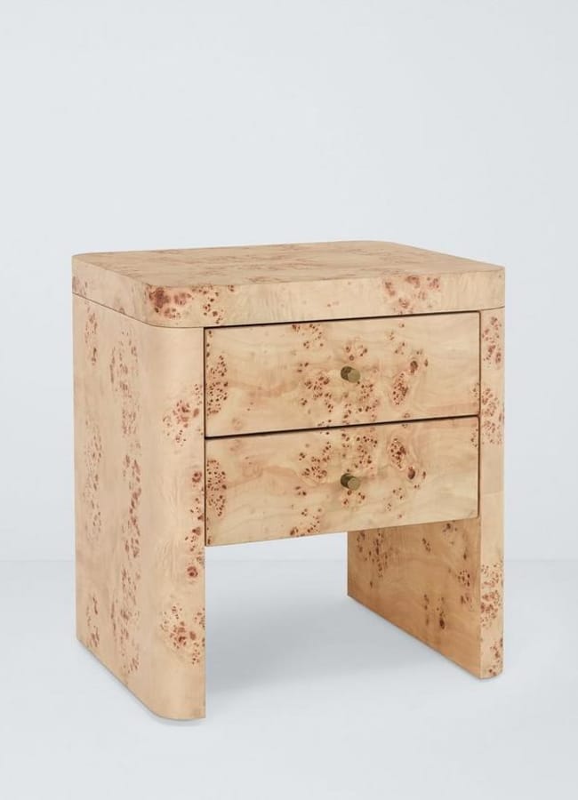A modern bedside table with a light wood finish, featuring two drawers with small round brass knobs and subtle natural wood grain patterns. The design has clean lines and a minimalist aesthetic.