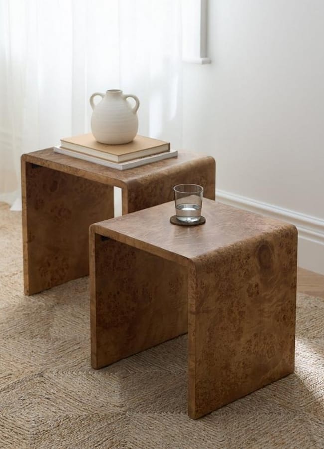 Two modern burr wood side tables with a curved design rest on a woven rug; one holds a glass of water, the other a stack of books and a ceramic vase, all bathed in soft natural light.