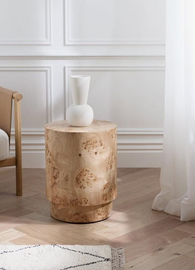 A round wooden side table with a light natural finish sits on a hardwood floor next to a beige chair. A white ribbed ceramic vase is placed on top, near a window with sheer white curtains.