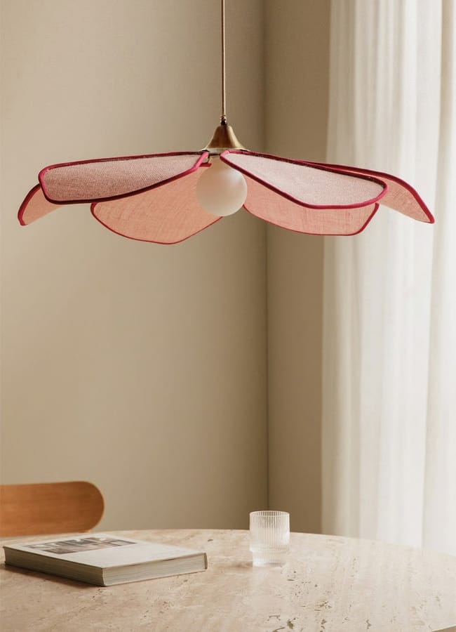 A pendant light with a pink, petal-shaped lampshade hangs above a round marble table, adding charm to red and pink interiors. On the table are a closed book and textured glass, with a wooden chair and sheer curtains in the background.