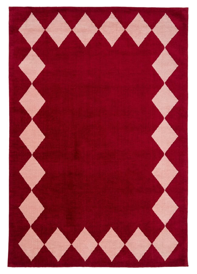 A rectangular red rug with a border of cream-coloured diamond shapes forms a geometric pattern around the edges. Ideal for red and pink interiors, its solid red centre adds warmth and elegance to any space.