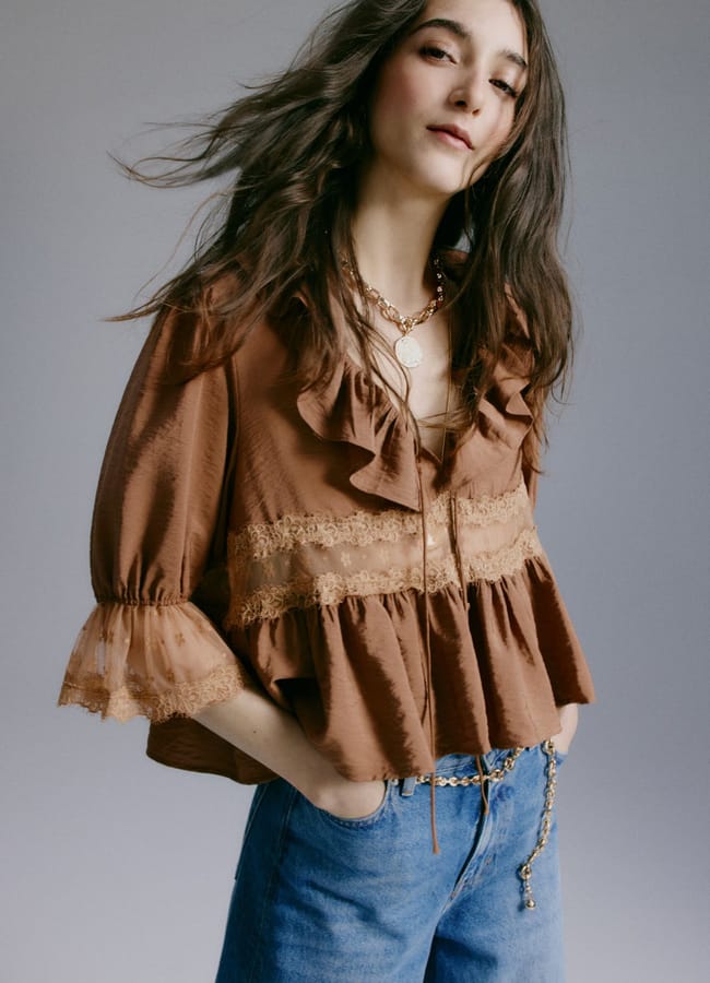 A woman with long brown hair wears a brown ruffled blouse with lace details and blue jeans from the H&M new season. She has her hands in her pockets and looks confidently at the camera against a plain background.