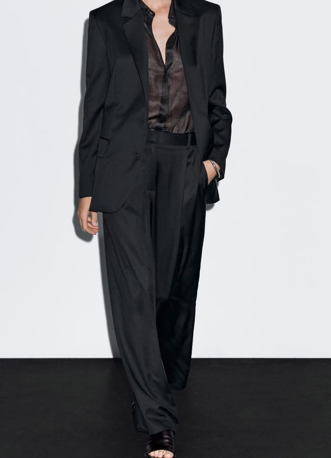 A person wearing a loose-fitting black suit with wide-leg pleated trousers and an unbuttoned sheer black shirt stands against a plain white background. Their face is not fully visible.