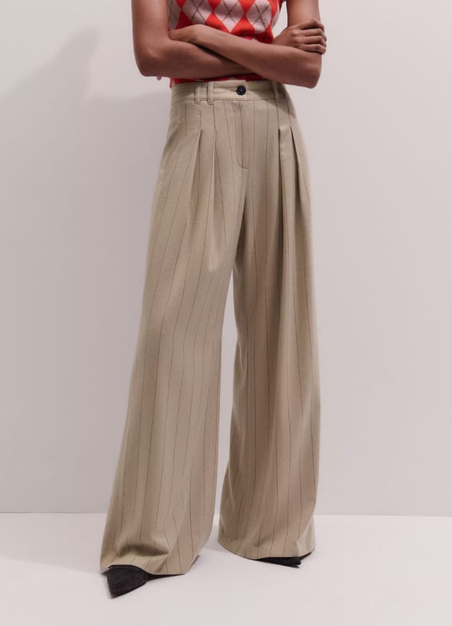 A person wearing high-waisted, wide-leg pleated trousers with vertical pinstripes, paired with a sleeveless red and grey argyle knitted top, stands against a plain background.