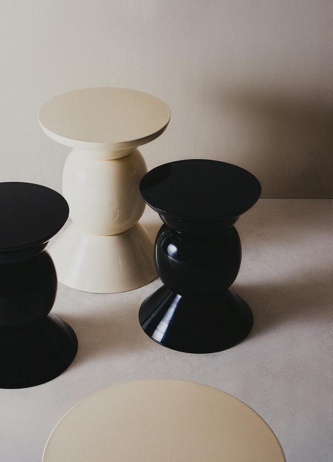 Four modern, round side tables in black and cream are arranged on a light surface against a neutral wall. Each table features a glossy finish and a sculptural, double-cone base—perfect for elevating any high street homeware collection.