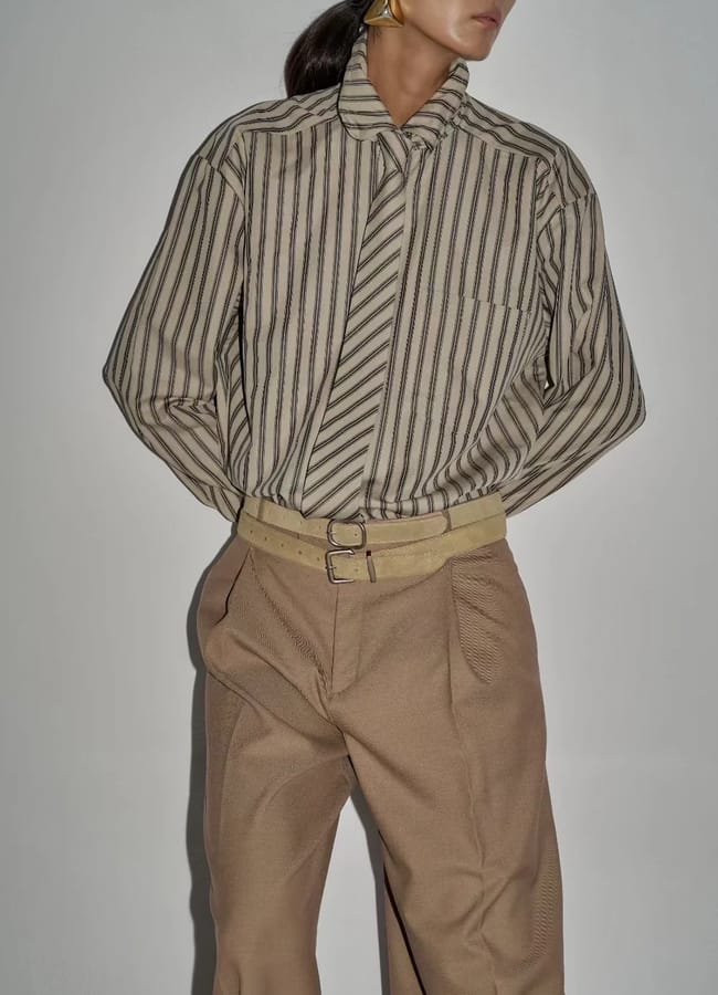 A woman wearing a tan-striped button-up shirt with a matching striped tie, beige high-waisted trousers, a beige belt, and geometric gold earrings stands against a plain white background, channelling a subtle Wuthering Heights wardrobe vibe. Her hands are behind her back.