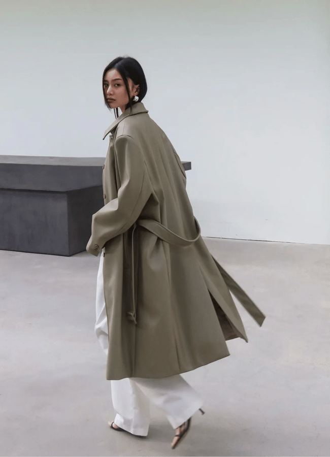 A woman wearing a long, loose, beige mac over white trousers and heeled sandals walks indoors, glancing back over her shoulder—a scene that channels a Wuthering Heights wardrobe vibe against minimal grey benches and blank walls.