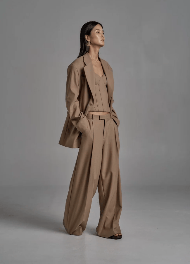A woman stands in a studio wearing an oversized brown trouser suit with pleated wide-leg trousers and a matching blazer over a fitted top. She has long dark hair, gold earrings, and an elegant, confident pose against a grey background.