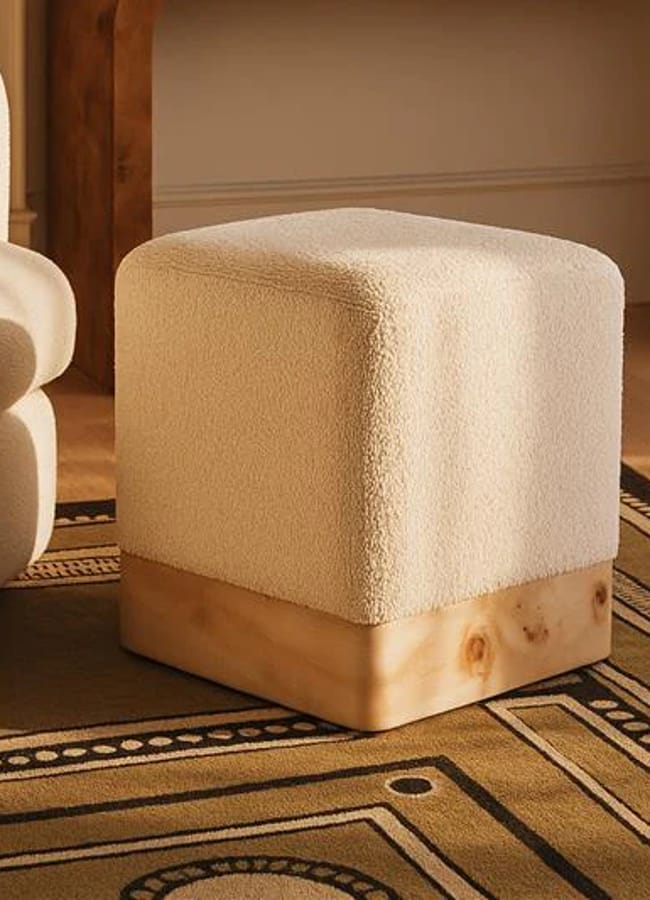 A modern, square ottoman with a textured cream fabric top and a smooth light wood base sits on a patterned rug next to a beige sofa in a warmly lit room.