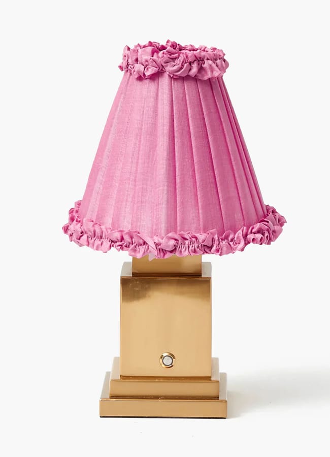 A gold square-based table lamp with a pleated pink lampshade featuring ruffled trim along the top and bottom edges, perfect for adding charm to red and pink interiors, set against a plain white background.