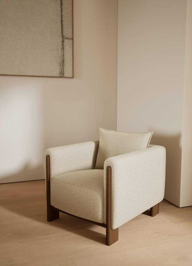 A modern beige armchair with wooden legs and armrests sits on a light wood floor in a minimalist room—an elegant piece inspired by high street homeware trends. A matching cushion rests on the chair, with abstract wall art in the background.