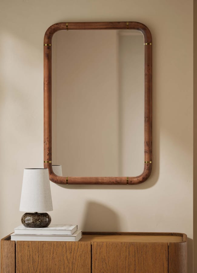 A rectangular mirror with a rounded wooden frame hangs on a beige wall above a wooden cupboard. A small table lamp with a white shade and stacked books sit on the cupboard.