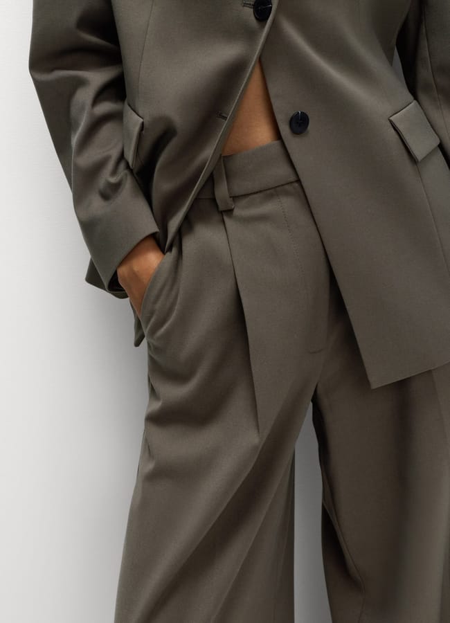 A person wearing a loose-fitting, olive green suit jacket and matching pleated trousers stands with a hand in their pocket, pictured from shoulders to thighs against a plain background.