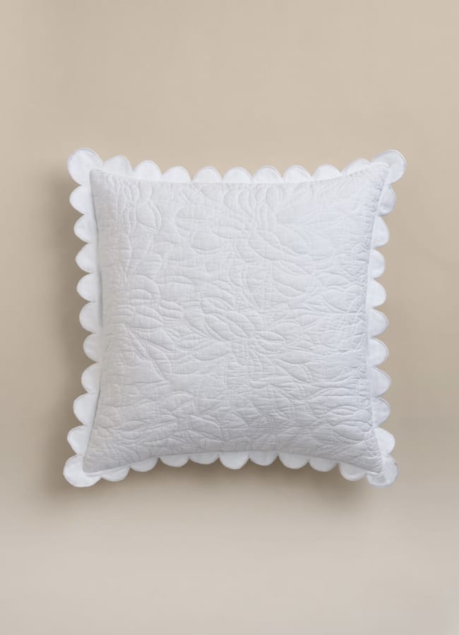 A white square cushion with a textured floral quilted pattern and scalloped edges, inspired by high street homeware trends, displayed against a neutral beige background.