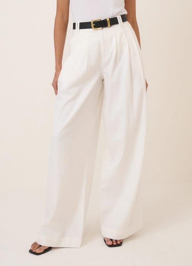 A person wearing wide-leg pleated trousers, a black belt with a gold buckle, a sleeveless white top, and black open-toe sandals stands against a plain light background. Only the lower half of the body is visible.
