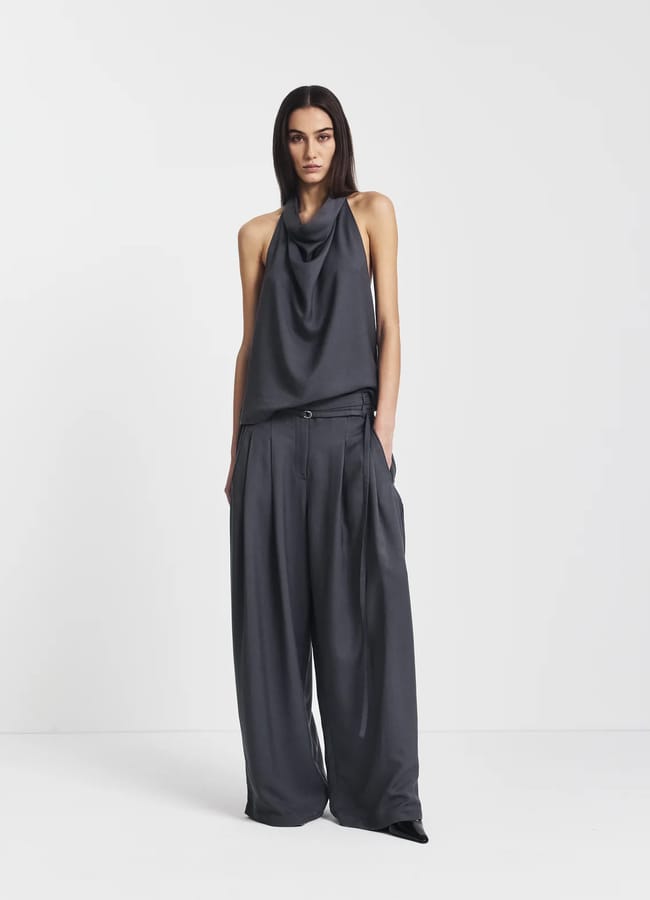 A woman stands against a plain white background, wearing a loose-fitting, dark grey halter top with a draped neckline and pleated trousers. Her hands are in her pockets and she has a neutral expression.