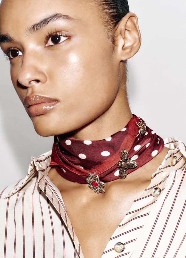 A person wearing a striped shirt with metallic buttons and a red scarf with white polka dots, adorned with ornate brooches as a fashion accessory, wrapped around their neck. Their hair is slicked back and their expression is neutral.