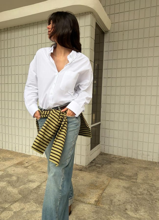A woman with shoulder-length dark hair wears a crisp white shirt, faded blue jeans, and has a striped jumper tied around her waist. She stands on a tiled pavement in front of a wall with square white tiles, looking to the side.