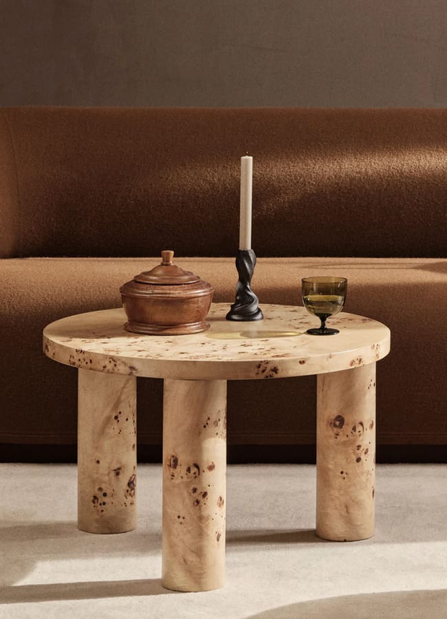 A round, three-legged wooden coffee table holds a brown lidded pot, a twisted black candlestick with a white candle, and a green glass goblet. A brown upholstered settee sits in the background.