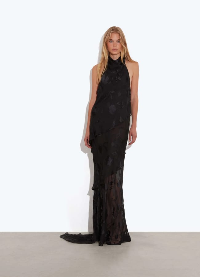 A woman reminiscent of Florence Pugh stands against a plain light background, wearing a sleeveless, floor-length black halter dress with a sheer overlay and subtle floral patterns. She has long, straight blonde hair and a neutral expression.