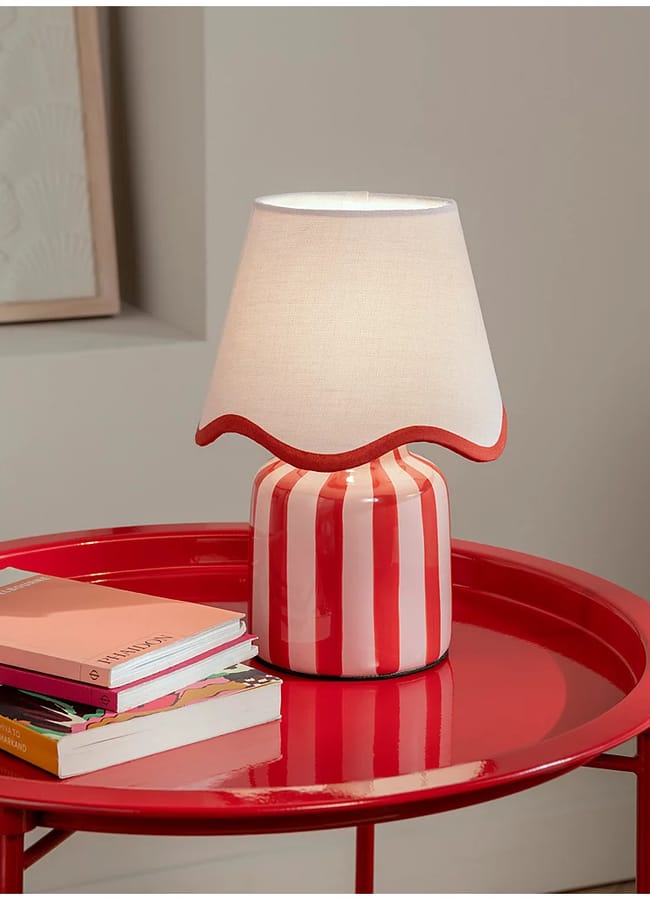 A red and white striped table lamp with a wavy-edged white lampshade sits on a glossy red round table, adding charm to red and pink interiors, next to a small stack of books against a neutral-coloured wall.