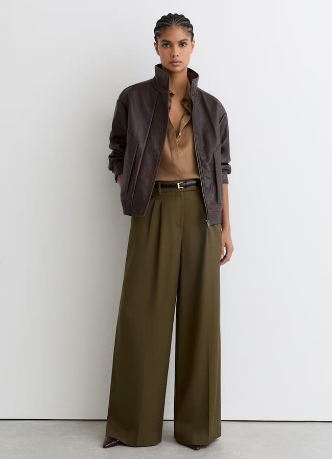 A woman stands against a white wall wearing a brown leather jacket, a tan blouse, wide-leg olive green pleated trousers, and brown pointed-toe shoes. Her hands are behind her back and her hair is styled in plaits.