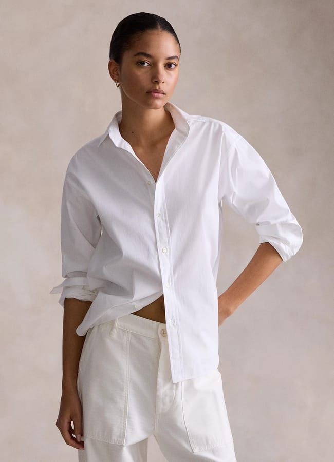 A person in a crisp white shirt with sleeves rolled up and white trousers stands against a neutral beige background, looking confidently at the camera. White shirts create an effortlessly classic and polished look.