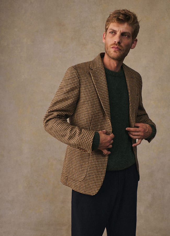 A man with light brown hair and a short beard wears a brown checked blazer over a dark green jumper and dark trousers—a perfect example of casual date night looks—while standing against a neutral textured background.