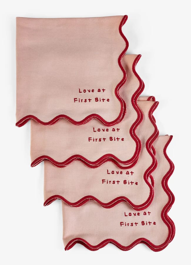 Four light pink cloth serviettes with red wavy edges, perfect for red and pink interiors, each embroidered with the phrase Love at First Bite in red lettering. The serviettes are neatly stacked and slightly overlapping.