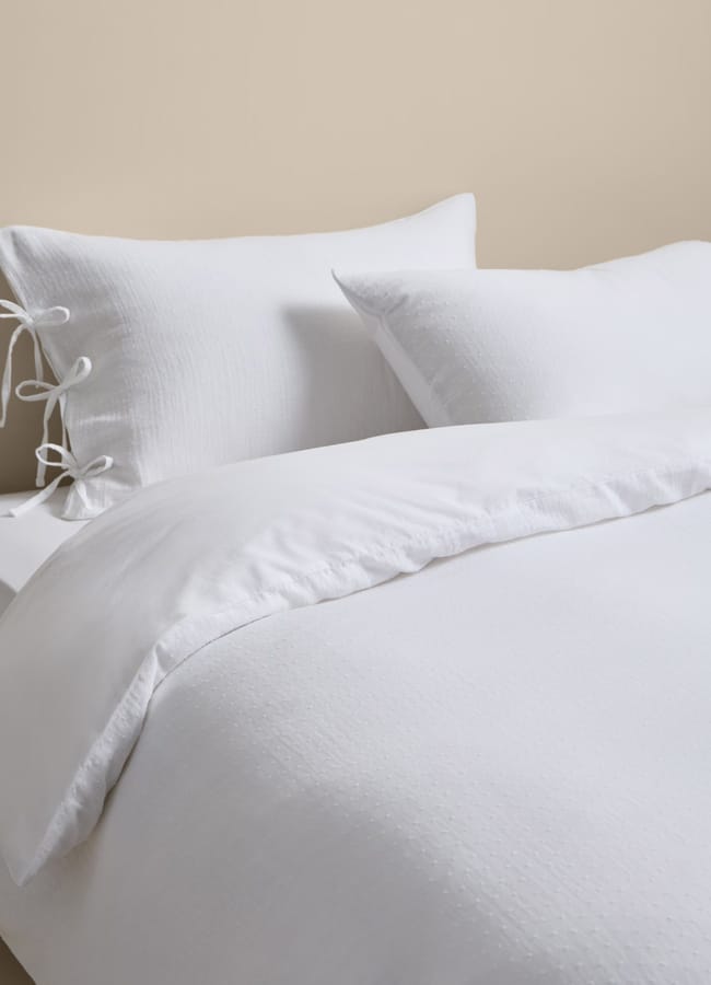 A neatly made bed with white textured bedding from high street homeware, two pillows—one with decorative side ties—against a beige wall, creating a clean and cosy appearance.