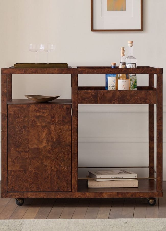 A wooden drinks trolley with a cupboard, shelves holding bottles and glasses, and a few magazines on the bottom shelf, stands against a white wall beneath a framed picture.