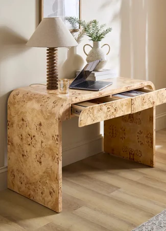 A modern light wood desk with curved edges, two open drawers, a lamp, a glass, and decorative items on top, placed against a beige wall on light wood flooring.