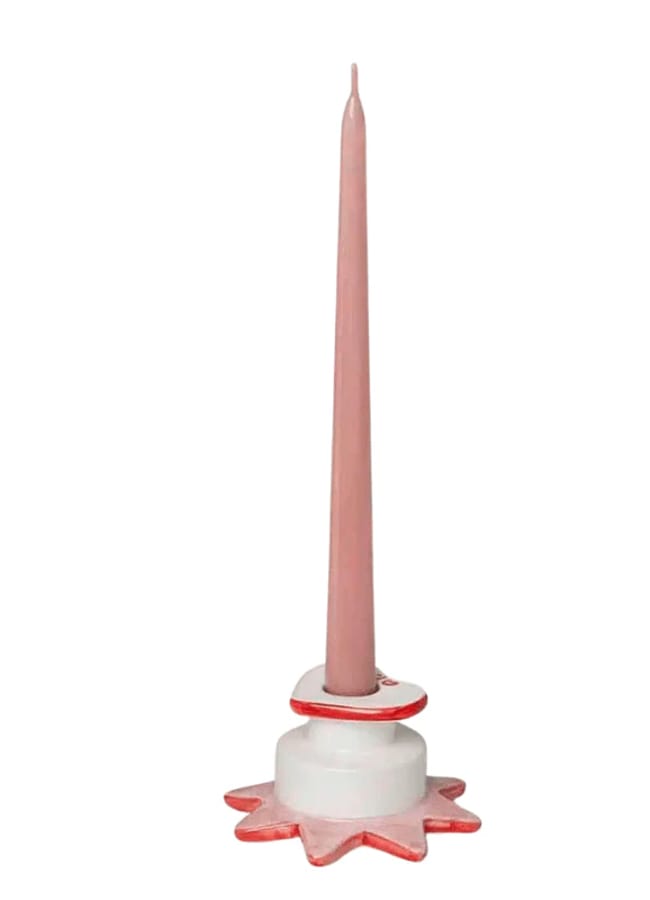 A tall, pink taper candle stands upright in a white ceramic holder with a pink-rimmed, star-shaped base—an elegant accent perfect for red and pink interiors. The holder and candle feature soft, pastel colours.