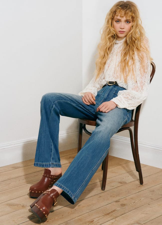 A person with long wavy blonde hair sits on a wooden chair in a corner, wearing an H&M new season white lace blouse, blue high-waisted jeans, and brown platform clogs with gold studs. The floor is light wood and the walls are white.