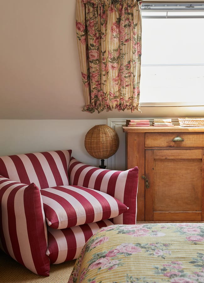 A cosy room with red and pink interiors features a red and white striped armchair, a wooden cupboard, a wicker globe lamp, floral curtains, and a floral bedspread. Natural light filters in from a window above the cupboard.