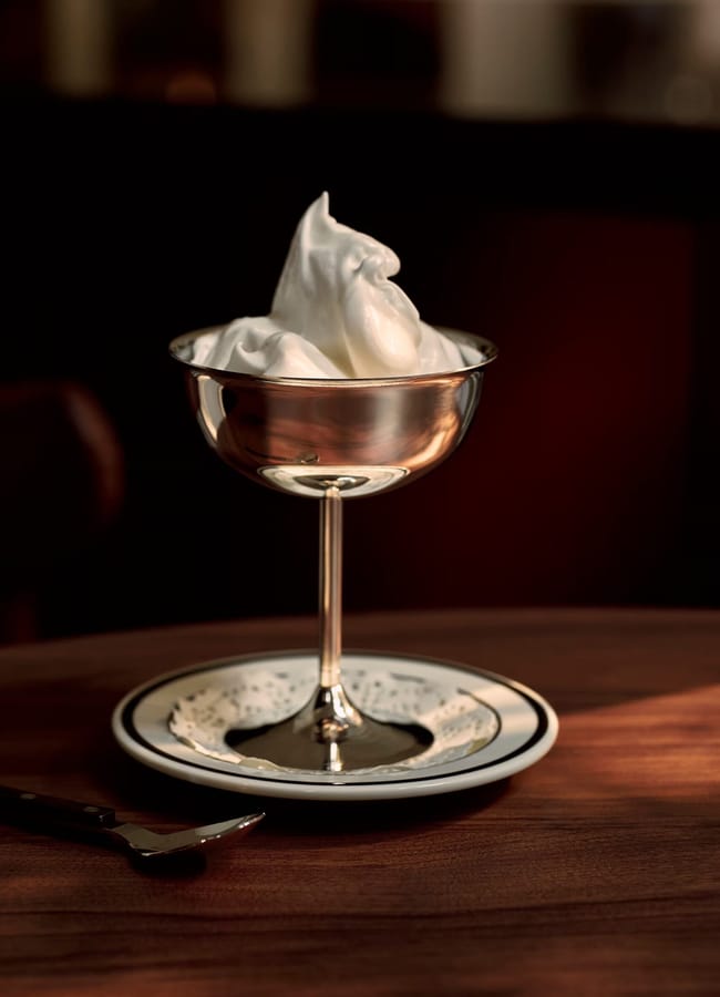 A silver coupe glass from high street homeware, filled with a swirl of whipped cream, sits on a small plate with a decorative paper doily, on a wooden table with a spoon beside it. Warm light creates a cosy atmosphere.