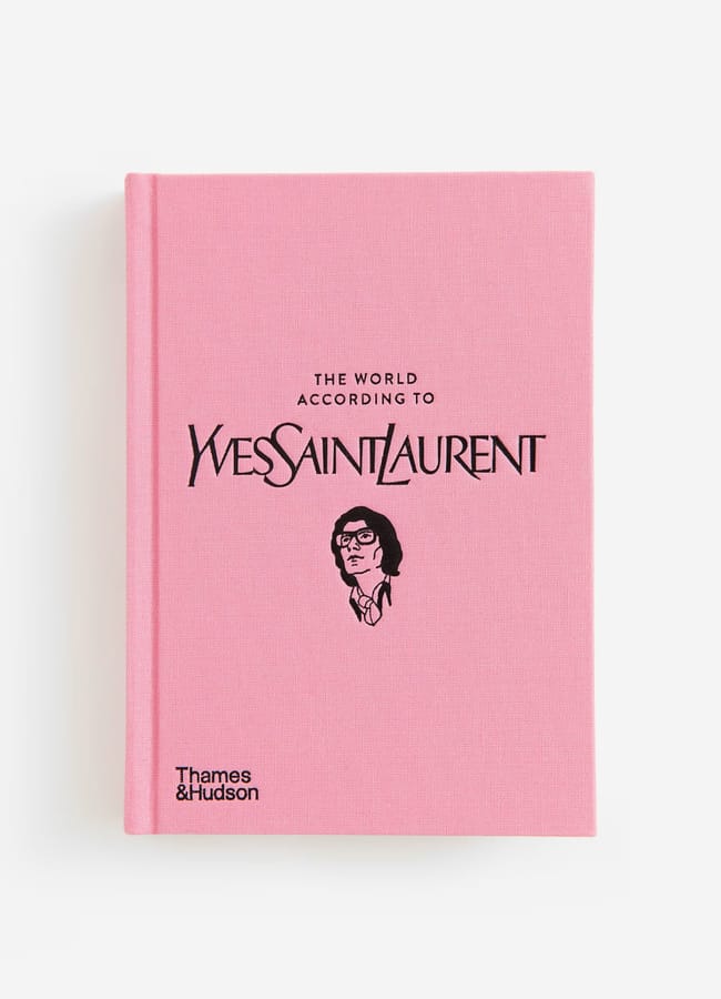 A pink hardback book titled The World According to Yves Saint Laurent with an illustrated portrait of a person wearing glasses, perfect for lovers of red and pink interiors, and Thames & Hudson written in the bottom left corner.