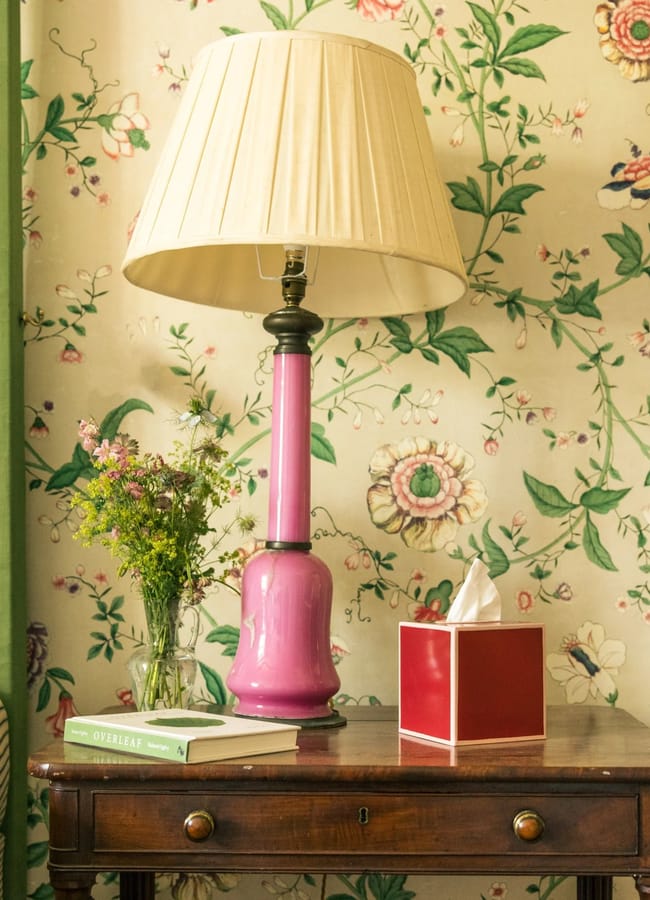A pink lamp with a cream shade sits on a wooden table beside a red tissue box, a glass vase with flowers, and books. The red and pink interiors are accented by floral wallpaper and a striped sofa partially visible on the left.
