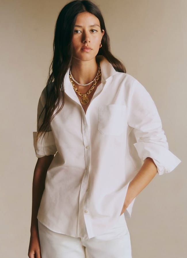 A woman with long dark hair wears a stylish white button-up shirt, showcasing the elegance of white shirts. She pairs it with white trousers and layered gold necklaces, standing confidently against a neutral background.