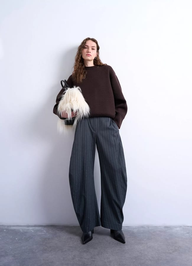 A woman stands against a plain wall, wearing a loose dark brown jumper and wide-leg, pleated grey trousers. She holds a fluffy white bag and wears pointed black shoes. Her long wavy hair frames her face as she gazes at the camera.