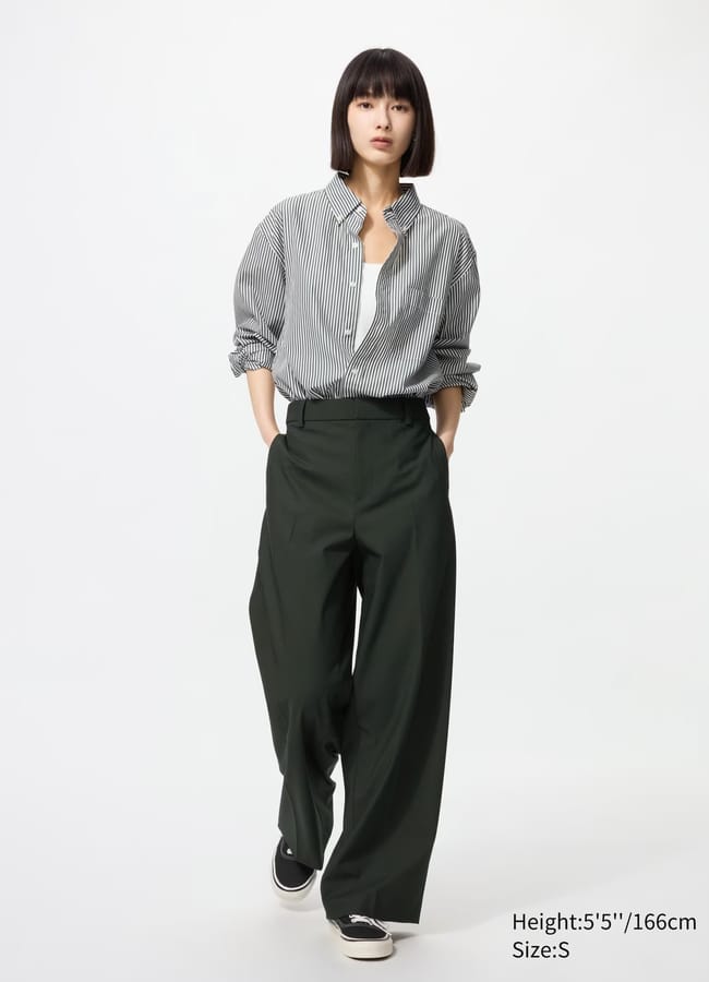 A person with straight dark hair wears a loose striped button-up shirt, dark pleated wide-leg trousers, and black slip-on shoes, standing against a plain white background. Text indicates height 5'5