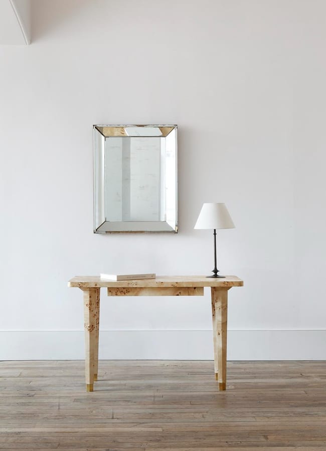 A minimalist room with a light wood table, a small black lamp with a white shade, a closed book, and a rectangular wall mirror above the table, set against a plain white wall and wooden floor.