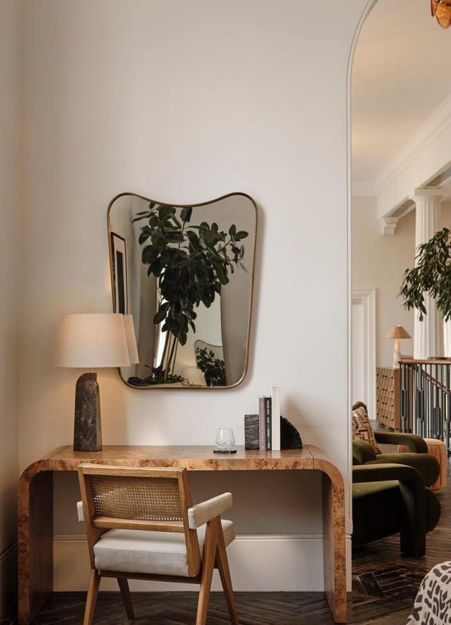 A small wooden desk with a lamp, books, and a chair sits against a white wall beneath an irregularly shaped mirror reflecting a leafy plant. The space opens to a bright, stylish living area with greenery.